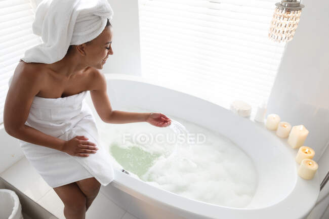 Улыбающаяся африканская американка, сидящая на краю ванны в ванной. оставаться дома в изоляции в карантинной изоляции — стоковое фото