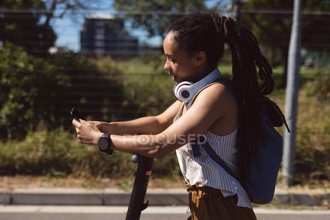 Afroamerikanerin mit Kopfhörer und Roller mit Smartphone auf der Straße. digitaler Nomade unterwegs. — Stockfoto