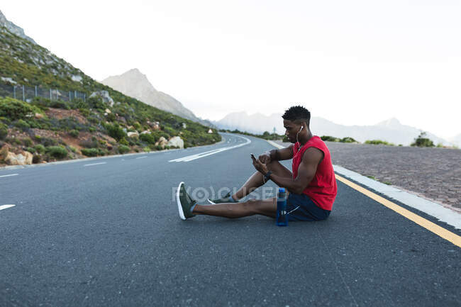 Hombre afroamericano haciendo ejercicio al aire libre usando un smartphone en una carretera costera. entrenamiento de fitness y estilo de vida saludable al aire libre. - foto de stock