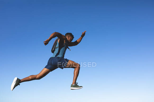 Hombre afroamericano haciendo ejercicio al aire libre saltando con el cielo azul en el fondo. entrenamiento de fitness y estilo de vida saludable al aire libre. - foto de stock