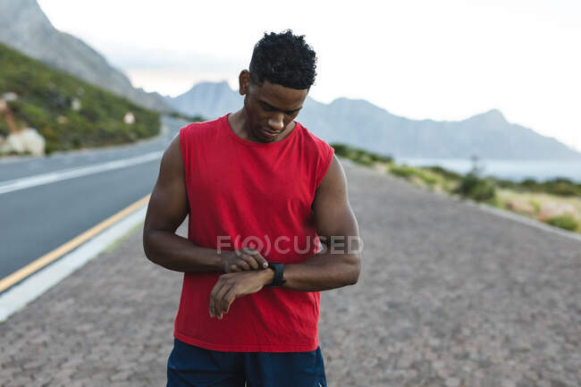Африканский американец тренируется на открытом воздухе, проверяя умные часы на прибрежной дороге. фитнес-тренировки и здоровый образ жизни. — стоковое фото