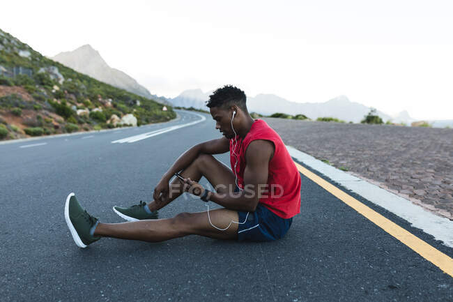Uomo afroamericano che si allena all'aperto usando lo smartphone su una strada costiera. allenamento fitness e stile di vita sano all'aperto. — Foto stock