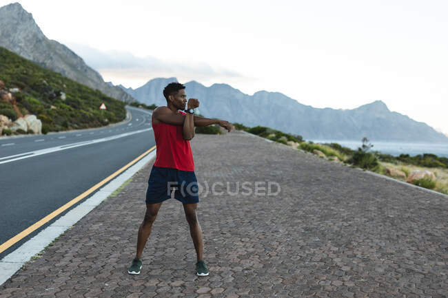 Hombre afroamericano ejercitándose al aire libre estirándose en una carretera costera. entrenamiento de fitness y estilo de vida saludable al aire libre. - foto de stock