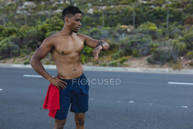 Hombre afroamericano haciendo ejercicio al aire libre revisando reloj inteligente en una carretera costera. entrenamiento de fitness y estilo de vida saludable al aire libre. - foto de stock
