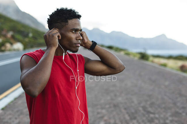 Hombre afroamericano haciendo ejercicio al aire libre poniendo auriculares en una carretera costera. entrenamiento de fitness y estilo de vida saludable al aire libre. - foto de stock