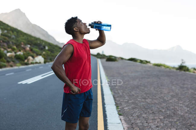 Homme afro-américain faisant de l'exercice en plein air de l'eau potable sur une route côtière. entraînement physique et mode de vie sain en plein air. — Photo de stock