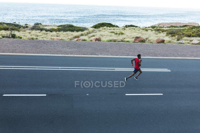 Африканский американец тренируется на открытом воздухе, бегая по прибрежной дороге. фитнес-тренировки и здоровый образ жизни. — стоковое фото