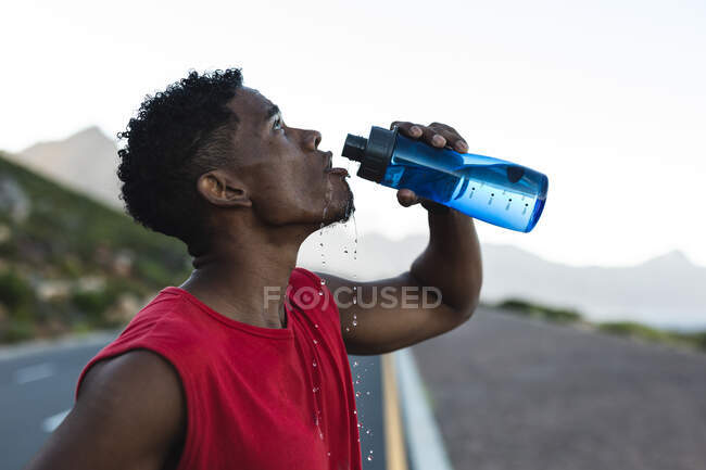 Hombre afroamericano haciendo ejercicio al aire libre bebiendo agua en una carretera costera. entrenamiento de fitness y estilo de vida saludable al aire libre. - foto de stock