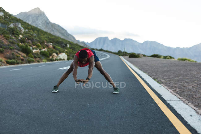 Homem afro-americano exercitando ao ar livre que se estende em uma estrada costeira. treinamento de fitness e estilo de vida saudável ao ar livre. — Fotografia de Stock