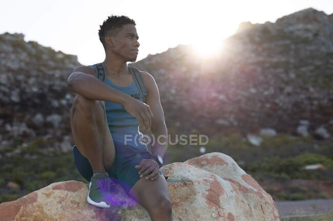 Hombre afroamericano haciendo ejercicio al aire libre sentado en una roca en una carretera costera. entrenamiento de fitness y estilo de vida saludable al aire libre. - foto de stock