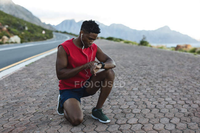 Hombre afroamericano haciendo ejercicio al aire libre revisando reloj inteligente en una carretera costera. entrenamiento de fitness y estilo de vida saludable al aire libre. - foto de stock