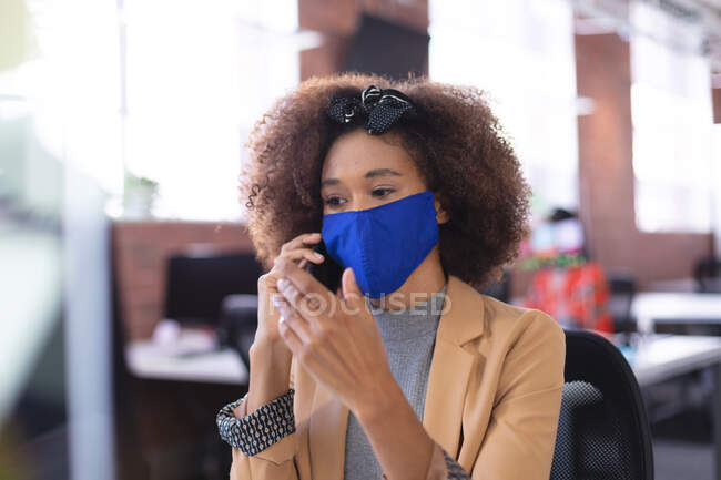 Африканская американка в маске разговаривает по смартфону в офисе. независимый бизнес креативного дизайна во время пандемии коронавируса ковида 19. — стоковое фото