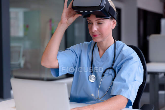 Médico femenino caucásico usando uniformes usando portátil y levantando auriculares vr. profesional médico en el trabajo con tecnología. - foto de stock