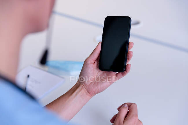 Over shoulder view of caucasian female doctor at desk using smartphone for video call consultation. telemedicine during quarantine lockdown. — Stock Photo