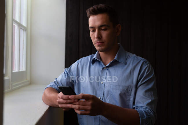Portrait of stylish caucasian businessman standing by window using smartphone. business person at work in modern office. — Stock Photo