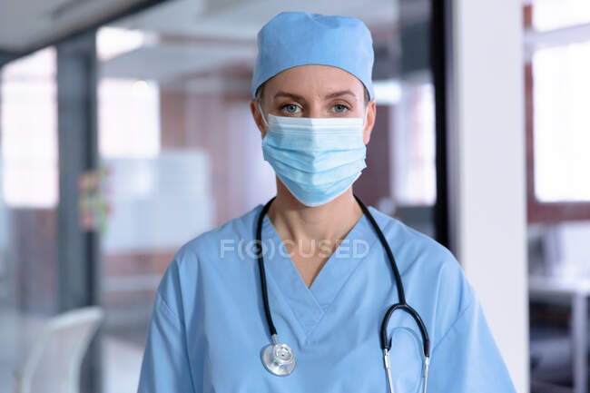 Retrato de doctora caucásica usando mascarilla facial, matorrales y estetoscopio. profesional médico en el trabajo durante la pandemia del coronavirus covid 19. - foto de stock