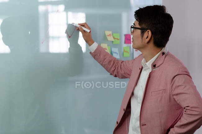 Stilvoller asiatischer Geschäftsmann schreibt Notizen an Glaswand. Geschäftsmann bei der Arbeit im modernen Büro. — Stockfoto