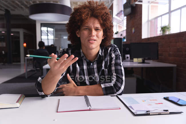 Empresario de raza mixta sentado en el escritorio haciendo gestos sosteniendo lápiz teniendo videollamada. negocio independiente de diseño creativo. - foto de stock
