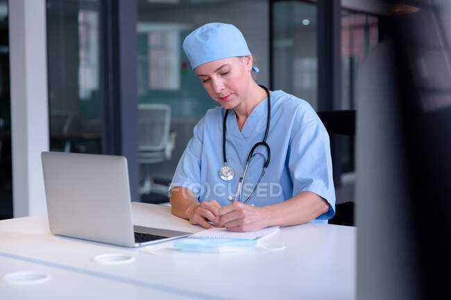 Médico femenino caucásico en el escritorio usando portátil tomando notas durante la consulta de videollamada. telemedicina durante el bloqueo de cuarentena. - foto de stock