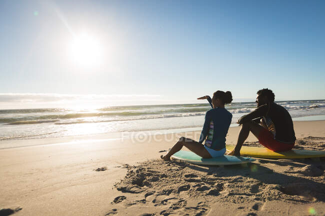 Glückliches afrikanisch-amerikanisches Paar am Strand, auf Surfbrettern sitzend und Richtung Meer blickend. gesunde Freizeit im Freien am Meer. — Stockfoto