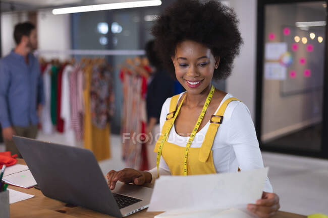 Stilista donna americana indossando metro nastro sarto in possesso di documento utilizzando il computer portatile. attività indipendente di design creativo. — Foto stock