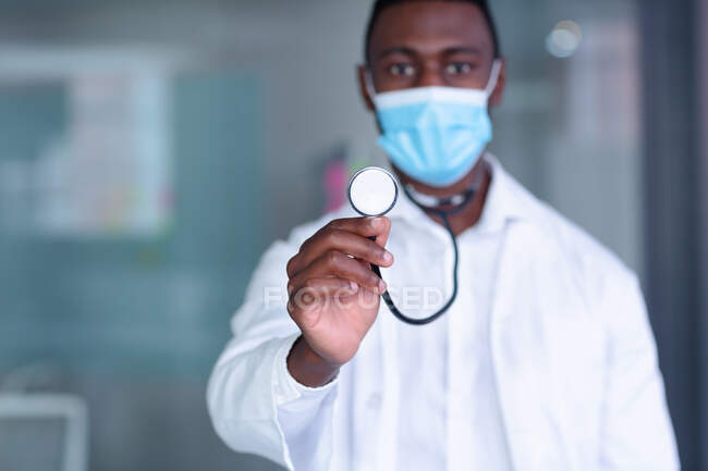 Портрет африканського лікаря чоловічої статі в масці, що тримає стетоскоп до камери. Медичний фахівець на роботі під час коронавірусної ковини 19 пандемії. — стокове фото