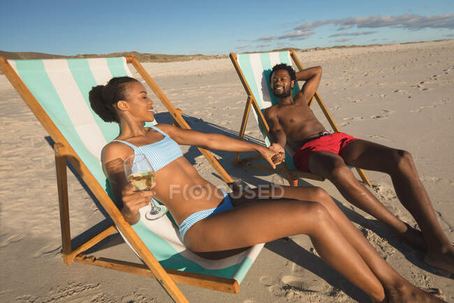 Pareja afroamericana enamorada sentada en tumbonas, cogida de la mano en la playa. amor, romance y vacaciones de verano de vacaciones de playa. - foto de stock