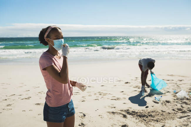 Afroamerikanisches Paar mit Mundschutz, das Müll am Strand sammelt. Öko-Strandschutz während der Coronavirus-Pandemie. — Stockfoto