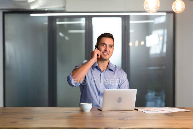 Porträt eines glücklichen kaukasischen Geschäftsmannes, der am Schreibtisch mit Laptop auf dem Smartphone spricht. Geschäftsmann bei der Arbeit im modernen Büro. — Stockfoto