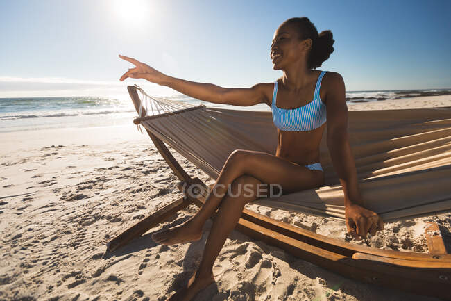 Mujer afroamericana feliz sentada en hamaca en la playa señalando. tiempo de ocio al aire libre saludable junto al mar. - foto de stock