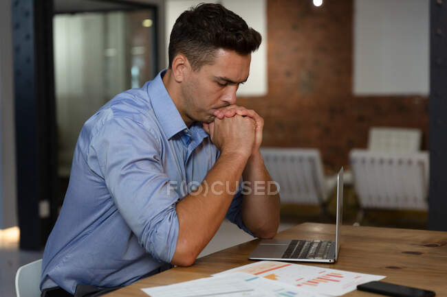 Portrait of stylish caucasian businessman thinking, sitting at desk and using laptop. business person at work in modern office. — Stock Photo