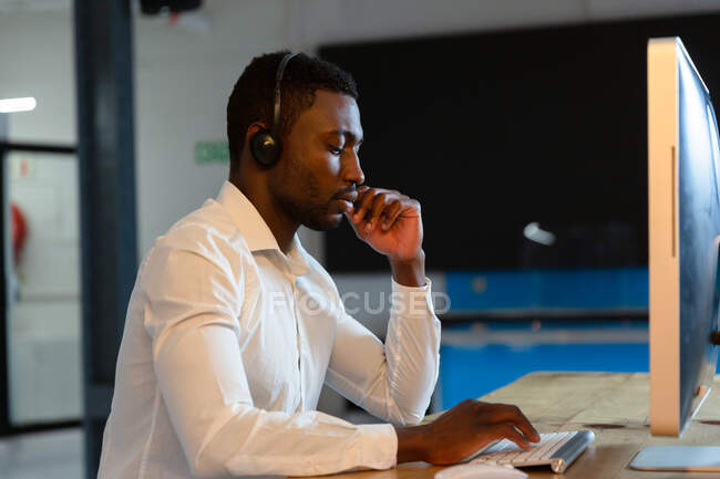 Uomo d'affari afroamericano casual che indossa cuffie telefoniche, usando il computer seduto alla scrivania. uomo d'affari al lavoro in ufficio moderno. — Foto stock