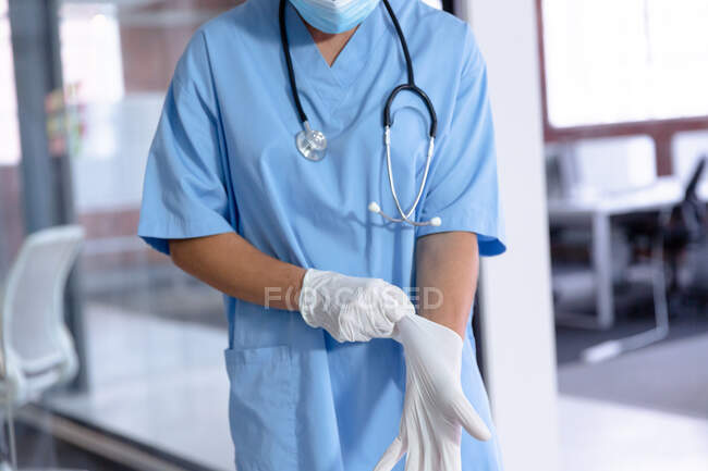 Midsection of caucasian female doctor wearing face mask and scrubs putting on surgical gloves. medical professional at work during coronavirus covid 19 pandemic. — Stock Photo