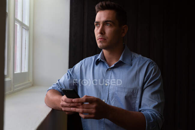 Portrait of stylish caucasian businessman thinking through window holding smartphone. business person at work in modern office. — Stock Photo