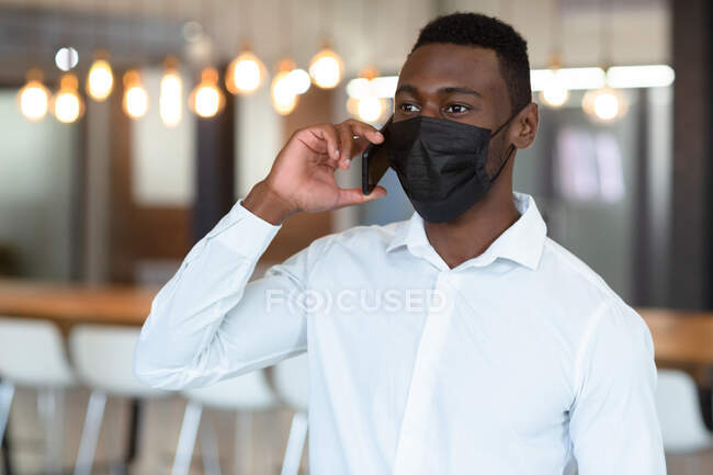 Lässiger afrikanisch-amerikanischer Geschäftsmann mit Gesichtsmaske, der auf dem Smartphone spricht. Geschäftsmann bei der Arbeit im modernen Büro während der Covid 19 Coronavirus-Pandemie. — Stockfoto