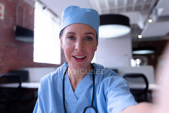 Doctora caucásica usando uniformes sentada en el escritorio hablando durante la consulta de videollamada. telemedicina durante el bloqueo de cuarentena. - foto de stock