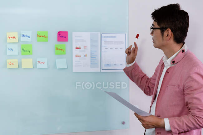 Stilvoller asiatischer Geschäftsmann beim Lesen von Zetteln, die ein Dokument mit Filzstift an einer Glaswand halten. Geschäftsmann bei der Arbeit im modernen Büro. — Stockfoto
