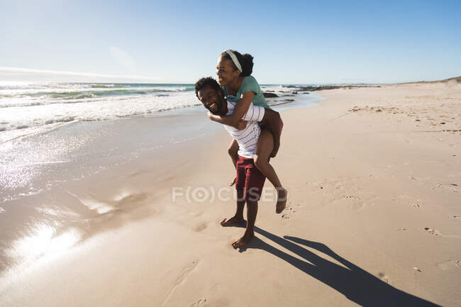 Happy african american couple on the beach piggybacking. healthy outdoor leisure time by the sea. — Stock Photo