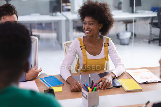 Feliz mujer de negocios afroamericana sentada en el escritorio en la sala de reuniones escuchando colegas. negocio independiente de diseño creativo. - foto de stock