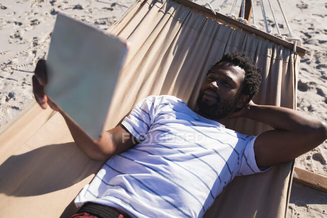 Homem americano africano feliz deitado em rede na praia usando tablet. tempo de lazer ao ar livre saudável pelo mar. — Fotografia de Stock