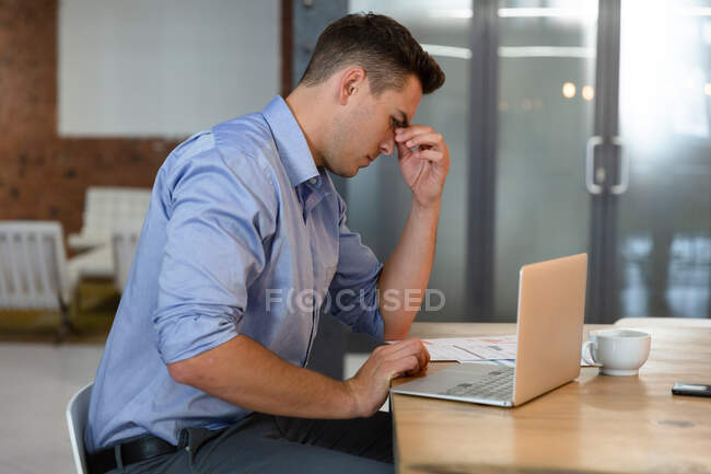 Casual hombre de negocios caucásico sentado en el escritorio, pensando y utilizando el ordenador portátil. persona de negocios en el trabajo en la oficina moderna. - foto de stock