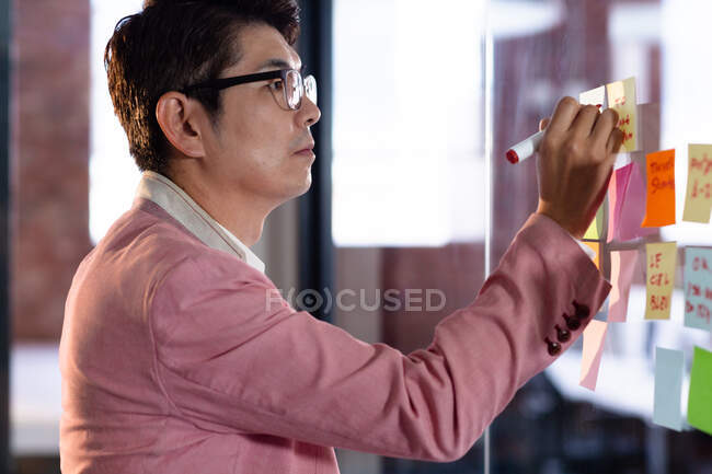 Стильний азіатський бізнесмен пише нотатки на скляній стіні. бізнесмен на роботі в сучасному офісі . — стокове фото