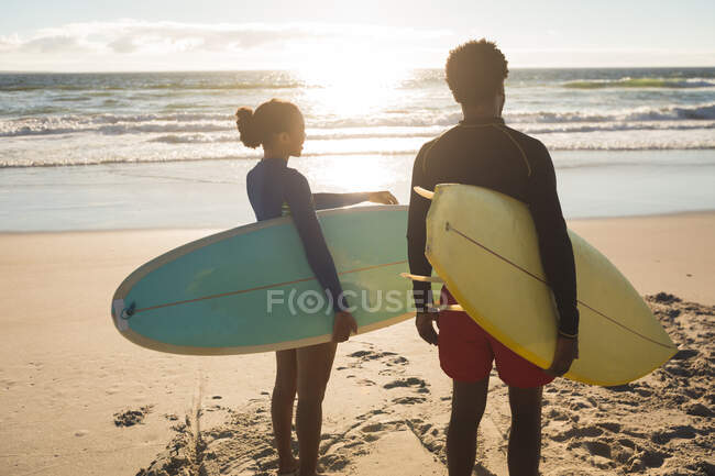 Felice coppia afroamericana sulla spiaggia portando tavole da surf. sano tempo libero all'aperto in riva al mare. — Foto stock