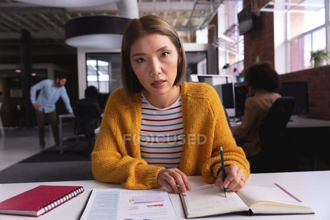 Donna d'affari di razza asiatica seduta alla scrivania a scrivere note con videochiamata. attività indipendente di design creativo. — Foto stock