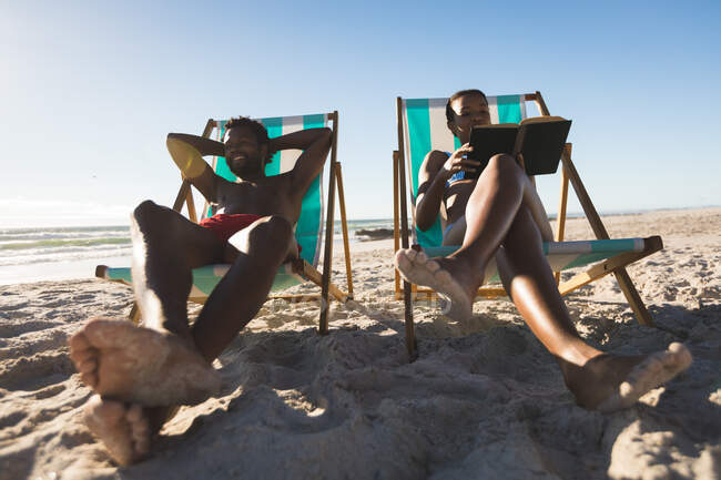 Casal afro-americano apaixonado sentado em cadeiras de praia, lendo livro relaxante na praia. amor, romance e férias de verão na praia. — Fotografia de Stock