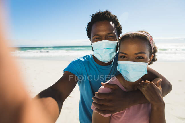 Ein afroamerikanisches Paar am Strand am Meer mit Masken, die ein Selfie machen. gesunde Freizeit am Meer während der Coronavirus-Pandemie 19. — Stockfoto