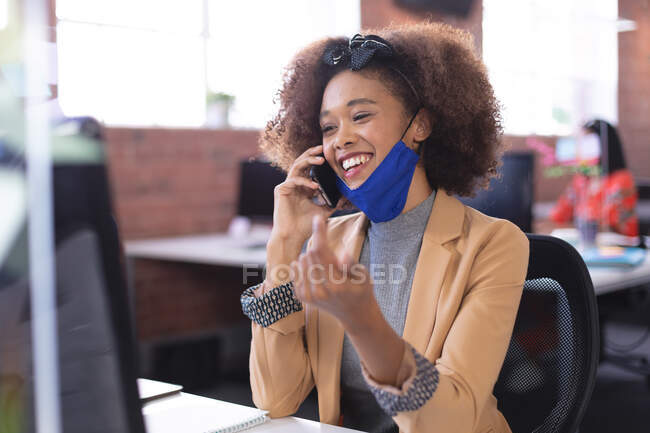 Feliz mujer de negocios afroamericana con máscara hablando por teléfono inteligente en la oficina. negocio independiente de diseño creativo durante la pandemia de coronavirus covid 19. - foto de stock