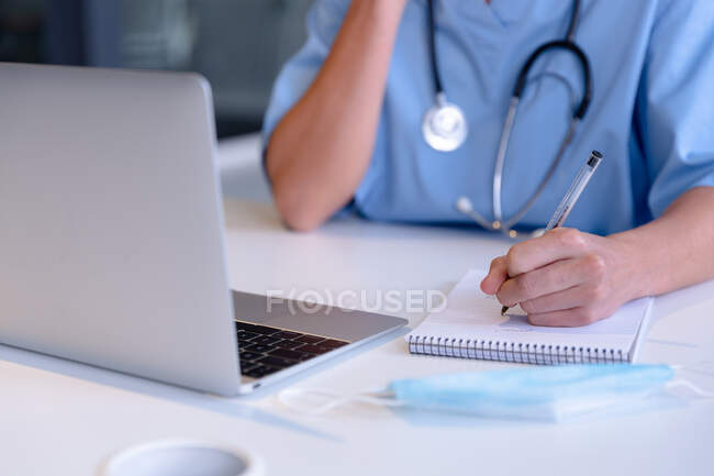 Midsection de médico feminino caucasiano usando laptop tomar notas durante a consulta de videochamada. telemedicina durante o bloqueio de quarentena. — Fotografia de Stock