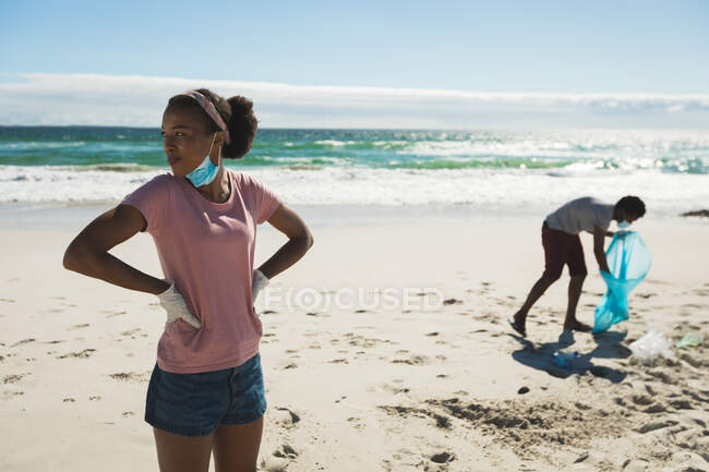 Pareja de raza afroamericana con mascarillas recogiendo basura de la playa. eco beach conservation during coronavirus covid 19 pandemic. - foto de stock
