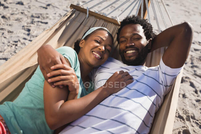 Happy african american couple lying in hammock on beach looking to camera. healthy outdoor leisure time by the sea. — Stock Photo